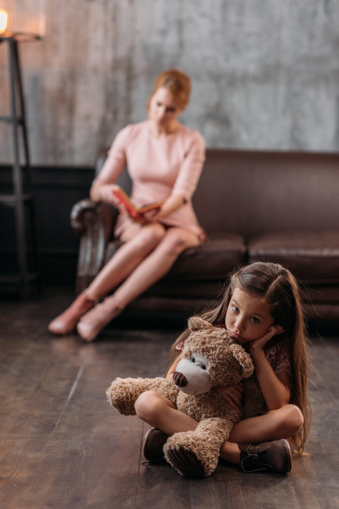 A little girl sitting on the floor with her stuffed animal, while her mother, who appears distant, sits on the couch in the background.