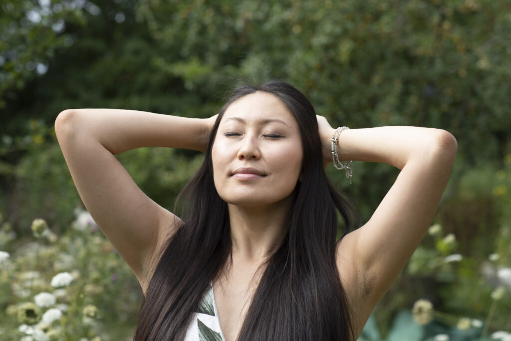 A mother in recovery stands outdoors with her hands on her head, smiling peacefully with her eyes closed as she practices mindfulness.